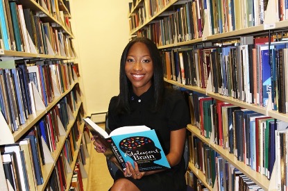 Brooke Preston sitting in between bookcases holding a book